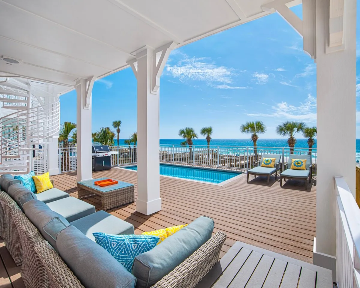 A vibrant exterior scene featuring a comfortable outdoor seating area beside a sparkling swimming pool, with a direct ocean view framed by palm trees. A built-in gas grill suggests the space is ready for barbecues and lively group gatherings. The mood is tranquil yet joyful, with clear cues for socializing and relaxation.