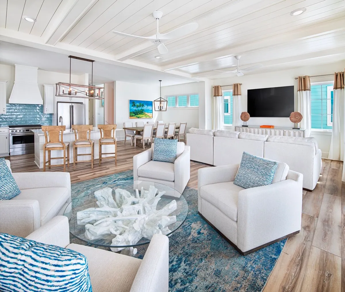 This image showcases a spacious, open-concept living area with white cushioned armchairs accented by blue patterned pillows, a glass coffee table with a coral centerpiece, and a view into the kitchen, which features a blue backsplash and wooden bar stools. The large flat-screen TV is prominently mounted on a white console, anchoring the living zone. The room is flooded with natural light, emphasizing the airy, inviting mood.