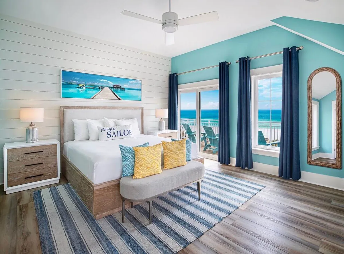 A serene king suite bedroom featuring a king-size bed with nautical-themed pillows, a large sliding glass door that opens to a private balcony with ocean views, a striped area rug, and an arched full-length mirror. The room exudes a calm, refreshing, and rejuvenating atmosphere, perfect for couples seeking privacy and luxury.
