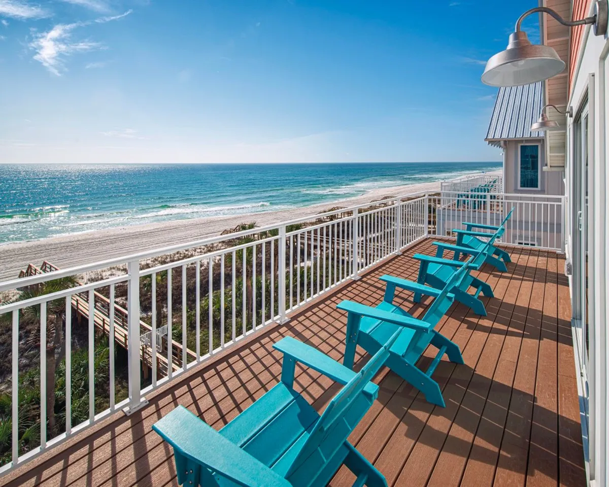A line of turquoise Adirondack chairs sits atop a spacious wooden balcony, framed by white railings. The balcony overlooks a stunning expanse of sandy beach and the sparkling blue Gulf, with a wooden dune walkover visible off to the side. The scene exudes tranquility and invites guests to enjoy morning coffee or evening sunsets while soaking up panoramic views.
