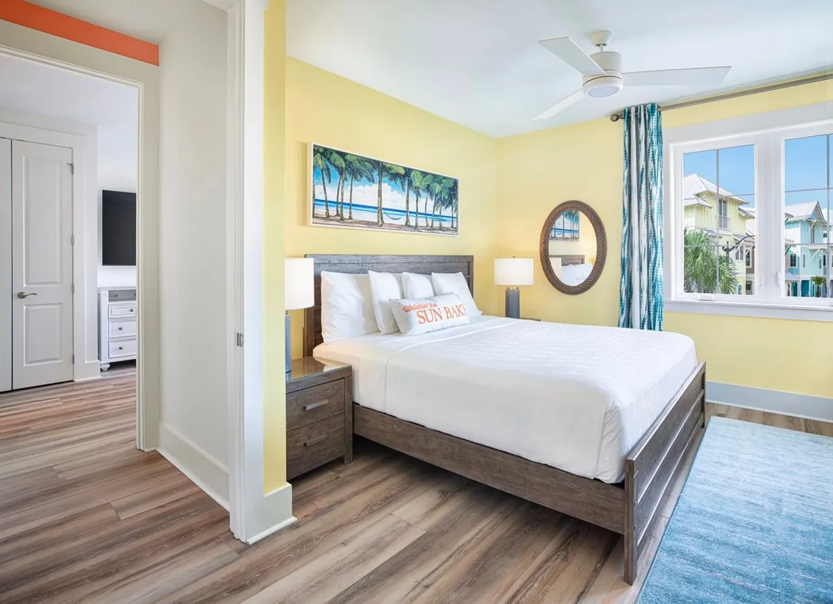 A bright, inviting bedroom featuring a wooden bed with crisp white linens, a cheerful yellow accent wall adorned with palm tree artwork, and a large window offering views of colorful beach cottages outside. The space is filled with natural light and decorated in a coastal style, evoking a serene vacation retreat.