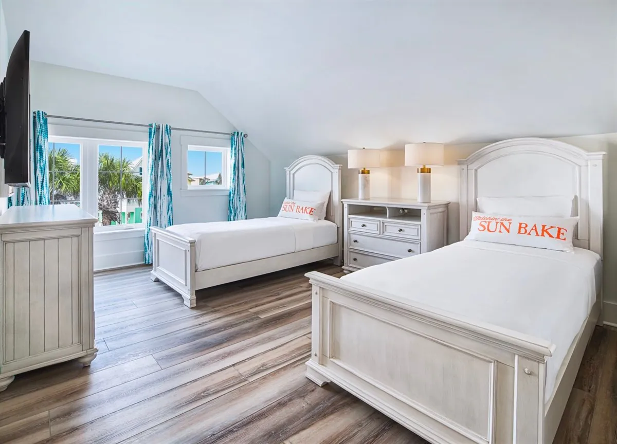 A bright and cheerful twin bedroom with two white wooden beds, white dresser and nightstand, blue and white patterned curtains, and large windows revealing palm trees outside. The room is airy and inviting, ideal for kids or teens, and offers flexible sleeping arrangements for families.