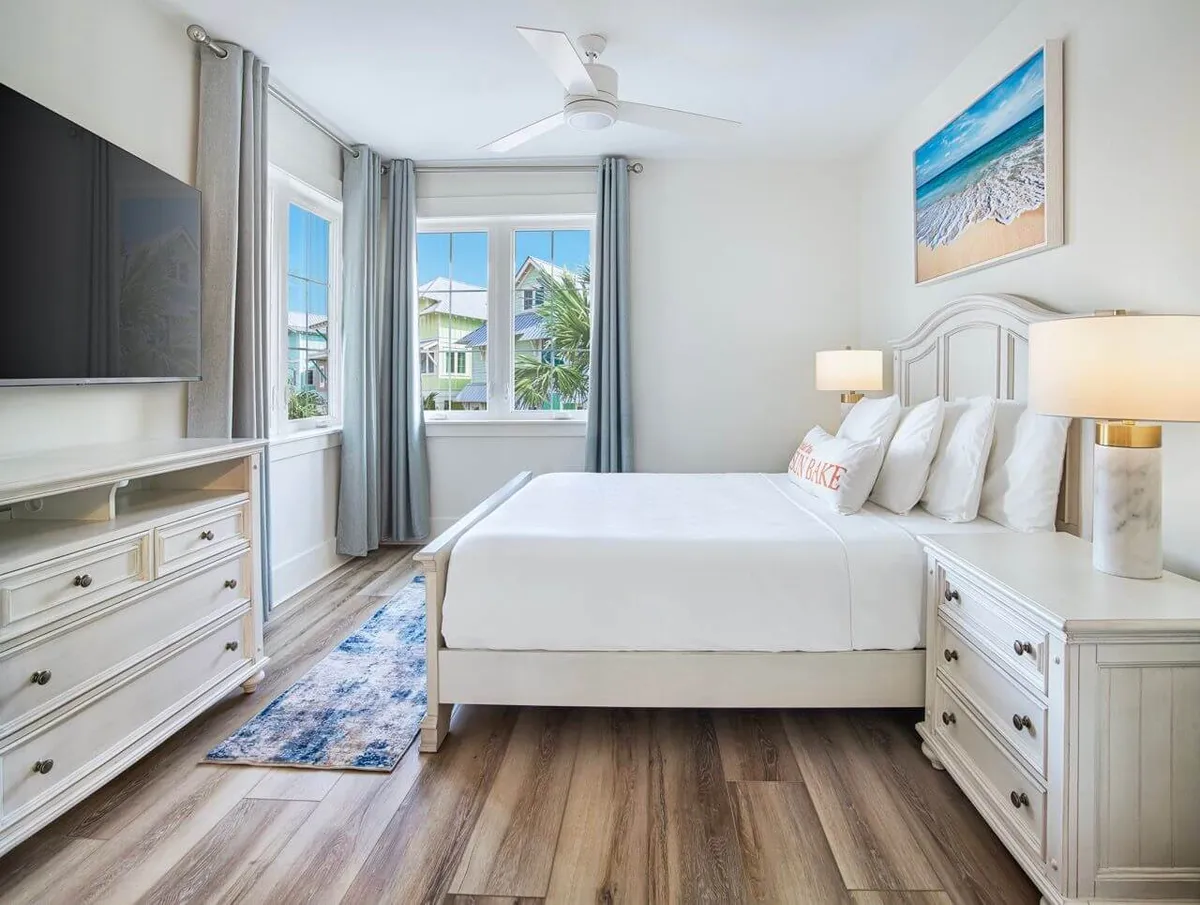 A peaceful bedroom with a white bed adorned with multiple pillows, large windows offering an ocean view, beach-themed wall art, and a wall-mounted flat screen TV. The space is tranquil and designed for restful, private moments.
