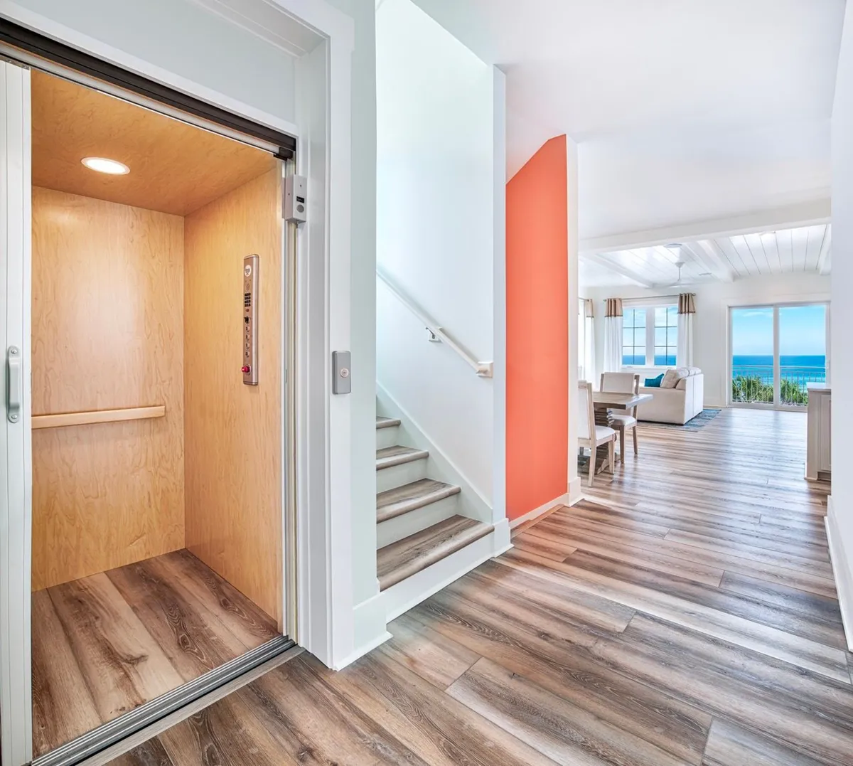 A bright, welcoming view of the living/dining area, featuring an open elevator with a wooden interior, a white-railed staircase, and a vibrant coral accent wall. Large windows in the living area frame a gorgeous Gulf view, and the space exudes accessibility and airy comfort.
