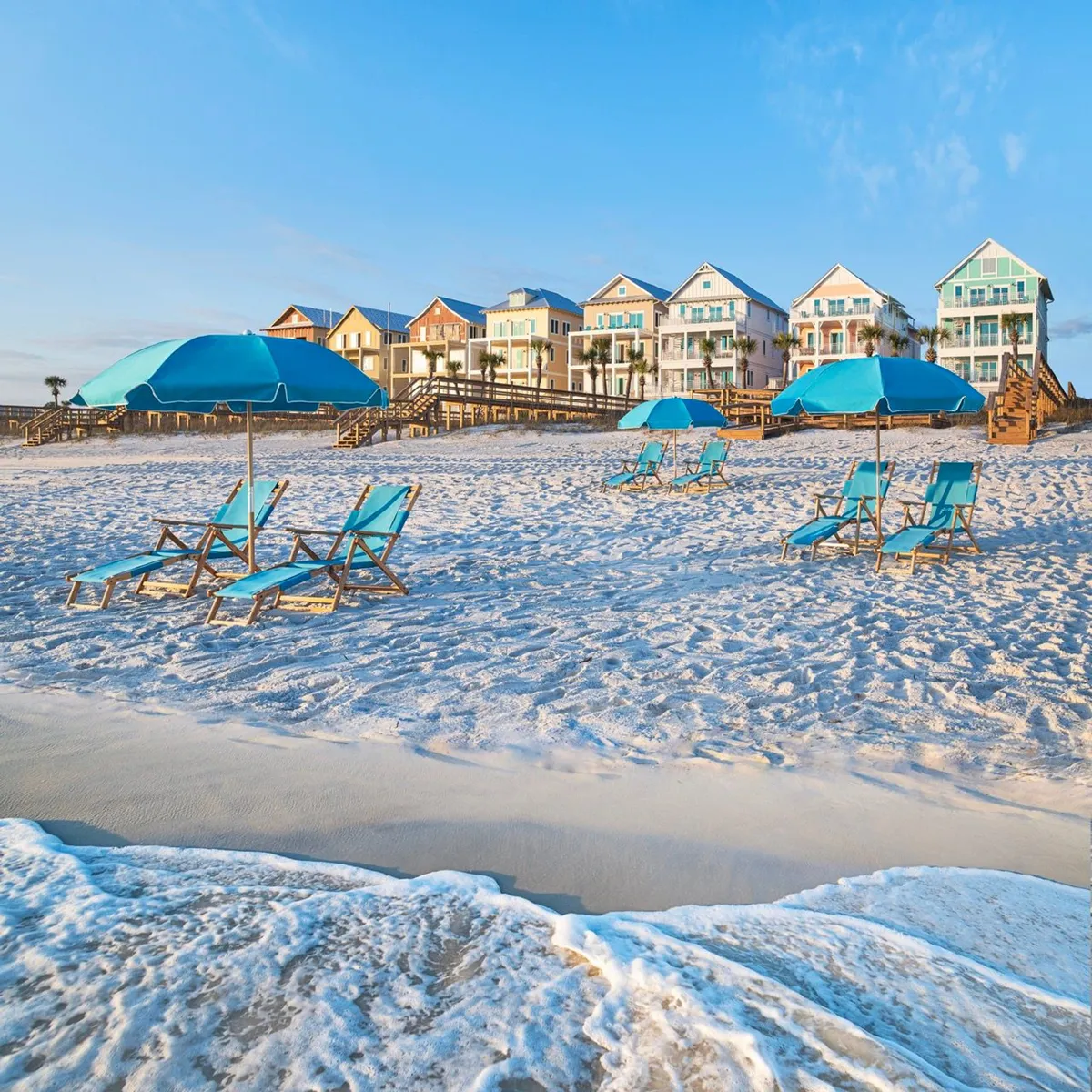 A vibrant row of colorful beachfront houses stretches along the sandy shore, each with multiple balconies offering panoramic Gulf views. Bright blue umbrellas and matching lounge chairs dot the beach, while wooden walkways connect the homes to the sand. Gentle ocean waves create a tranquil backdrop, and guests can be seen enjoying the sun, relaxing, and strolling along the pathways.