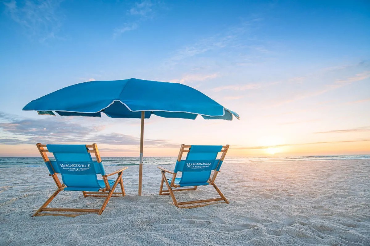 Two blue branded beach chairs and an umbrella are set up on the sandy shore, facing the calm Gulf waters. The sun sets on the horizon, casting golden light across the scene, creating a tranquil, idyllic beach atmosphere ideal for unwinding after a day of activities.