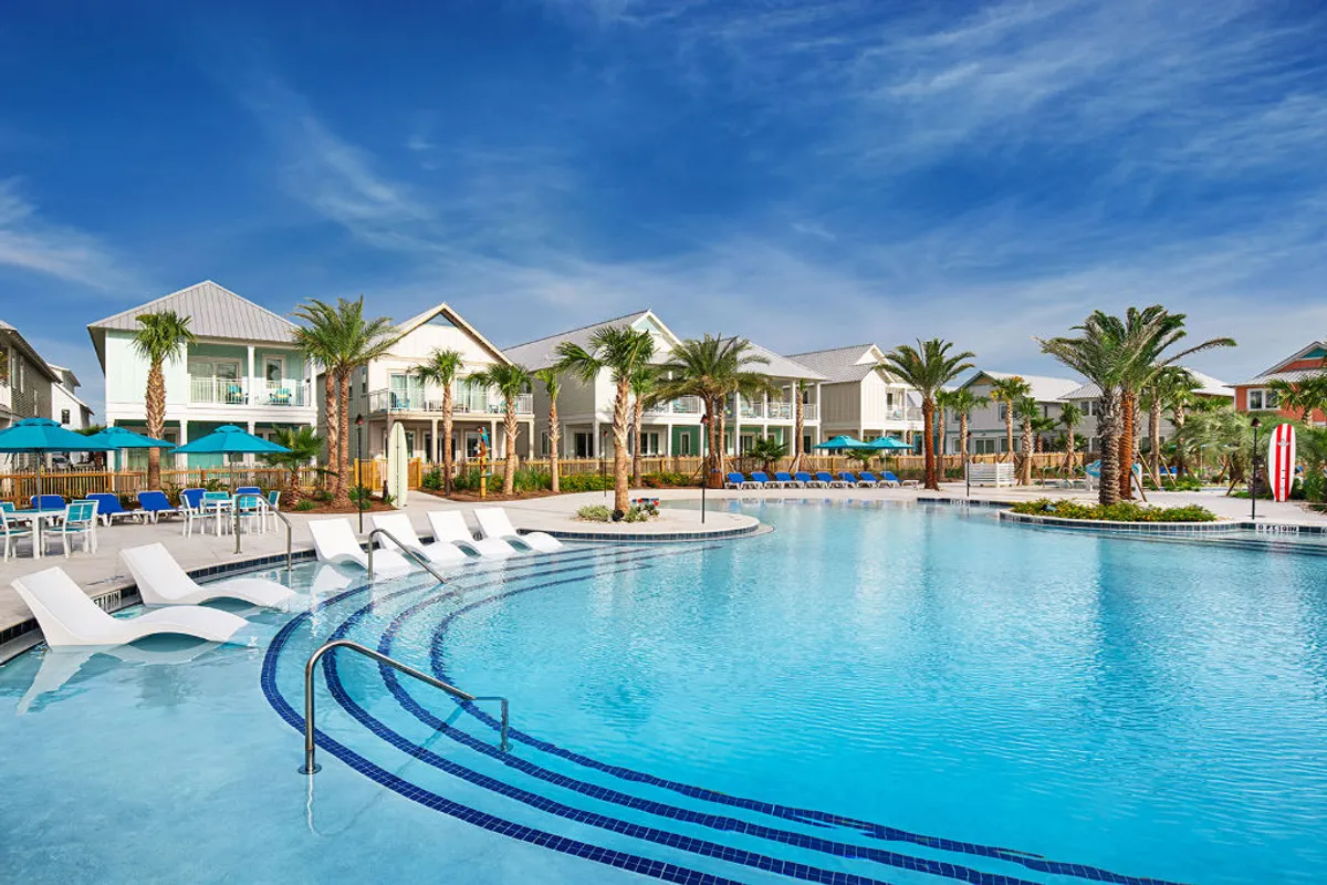 A large, heated outdoor pool surrounded by modern two-story beach cottages with balconies, tall palm trees, landscaped greenery, and ample poolside seating with umbrellas. The scene is lively yet relaxing, capturing guests swimming, lounging, and enjoying the resort amenities just steps from their villas.