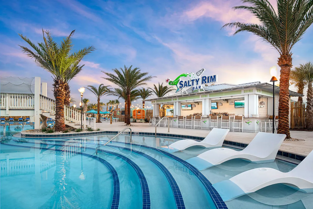 The lively Salty Rim Bar & Grill sits poolside, surrounded by palm trees and tropical landscaping. A large, curved pool with built-in lounge chairs submerged in the water invites guests to relax, swim, or socialize. The area buzzes with activity as families and friends enjoy poolside food and drinks, with the festive resort atmosphere apparent throughout.