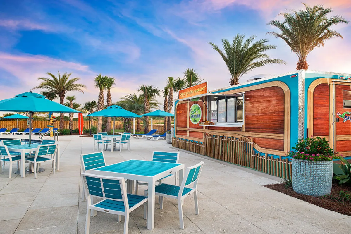 A festive poolside scene featuring turquoise and white outdoor tables and chairs shaded by blue umbrellas, surrounded by tropical palm trees. A lively wood-paneled outdoor bar with colorful signage is visible, inviting guests to enjoy drinks and snacks. Groups of people are relaxing, socializing, and dining in the warm, inviting atmosphere.