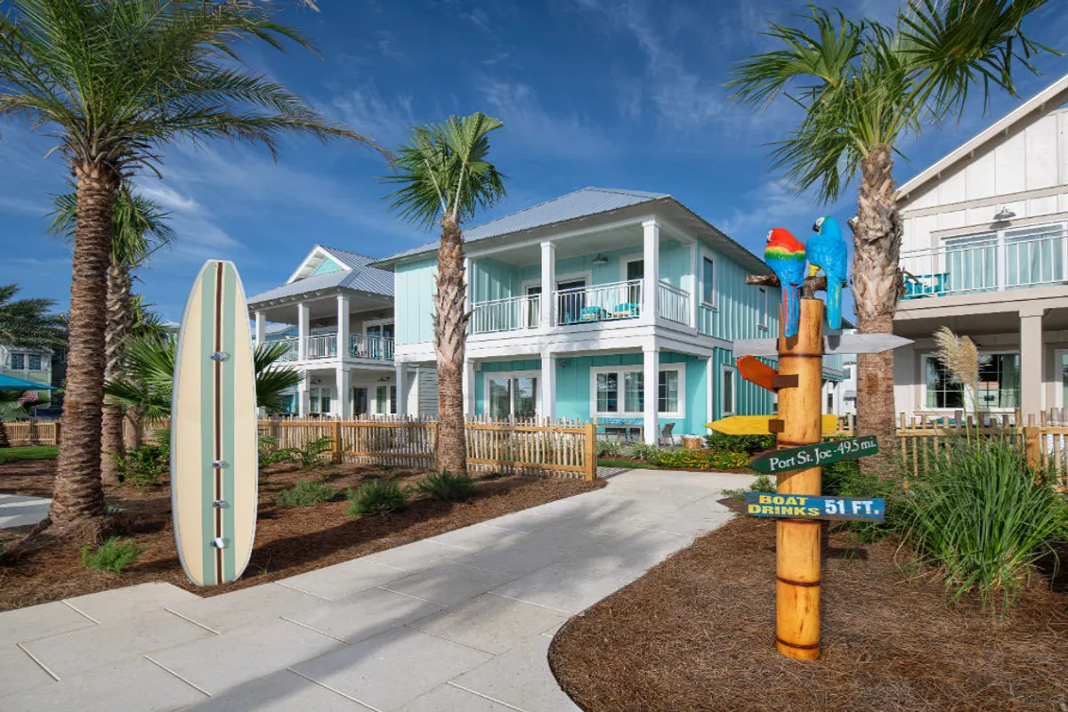 A cheerful exterior view of a turquoise beach villa with private balconies overlooking tropical landscaping. Decorative surfboards and a whimsical signpost with colorful parrots add a fun, coastal touch. Palm trees line the walkway, suggesting easy access to the beach and communal spaces for relaxation and group activities.