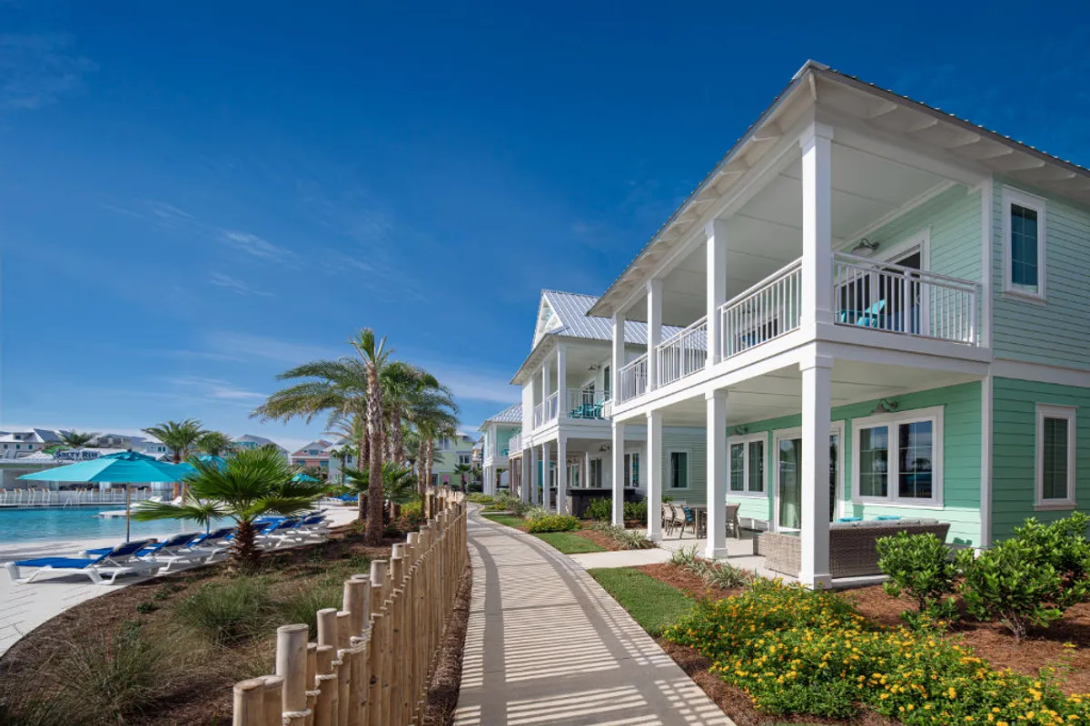 A pastel mint green beachfront villa with inviting poolside lounge chairs shaded by turquoise umbrellas. The patio and balconies are furnished for relaxation, and tropical landscaping with palm trees enhances the cheerful beach vibe.